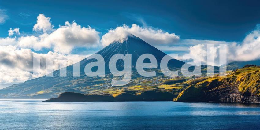 A majestic volcanic peak rises dramatically from the ocean, shrouded in clouds against a vibrant blue sky